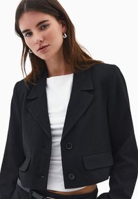 Blazer corto a righe nere con colletto a punta, due tasche frontali e chiusura a doppio bottone, indossato sopra una maglietta bianca.
