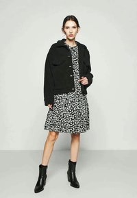 Casaco preto texturizado com botões prateados sobre um vestido preto com padrão floral. A modelo usa botas pretas até o tornozelo. Design simples e forma ajustada.