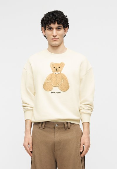 Νέος άνδρας με σγουρά μαλλιά, φοράει μια κρεμ φούτερ με 3D γραφικό αρκουδάκι και καφέ παντελόνι, σε απλό φόντο.