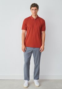 Polo rouge à manches courtes avec texture en diamants et col zippé, associé à un pantalon gris clair et des baskets blanches. Fond uni.
