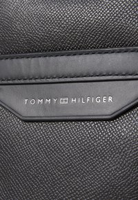 Material de piel negra texturizada con el logo "TOMMY HILFIGER" en plata. Costura detallada y una forma distintiva.