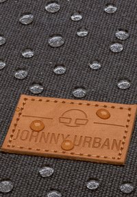 Szara tkanina z kroplami wody oraz brązowa skórzana metka z wytłoczonym napisem „JOHNNY URBAN”, zdobiona akcentami w postaci przeszyć i minimalistycznym designem.