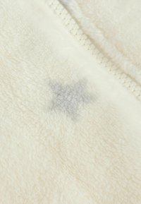 Krämfärgad fleece med en mjuk textur och en synlig grå stjärnformad fläck nära dragkedjedetaljen.