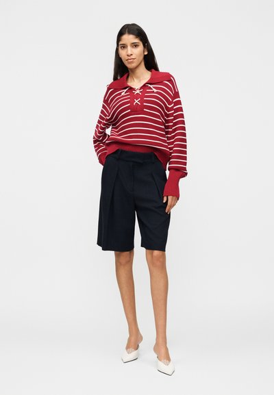 Γυναίκα όρθια, φοράει κόκκινο και λευκό ριγωτό πουλόβερ, μαύρες σορτς μέχρι το γόνατο και λευκά μυτερά παπούτσια μπροστά σε λευκό απλό φόντο.