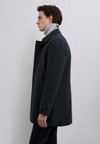 Abrigo de lana gris oscuro con un cuello alto, botones de doble botonadura y bolsillos laterales. Cuello de tortuga enrollado por debajo, diseño ajustado.