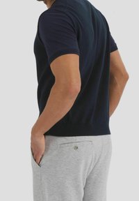 Uomo che indossa una camicia nera a maniche corte e pantaloni grigio chiaro, con una mano in tasca, visto da dietro su uno sfondo semplice.