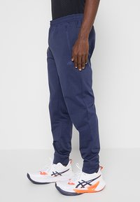 Granatowe spodenki sportowe z elastycznym paskiem, zwężającym się kształtem i teksturą materiału. Noszone z białymi i pomarańczowymi butami sportowymi.