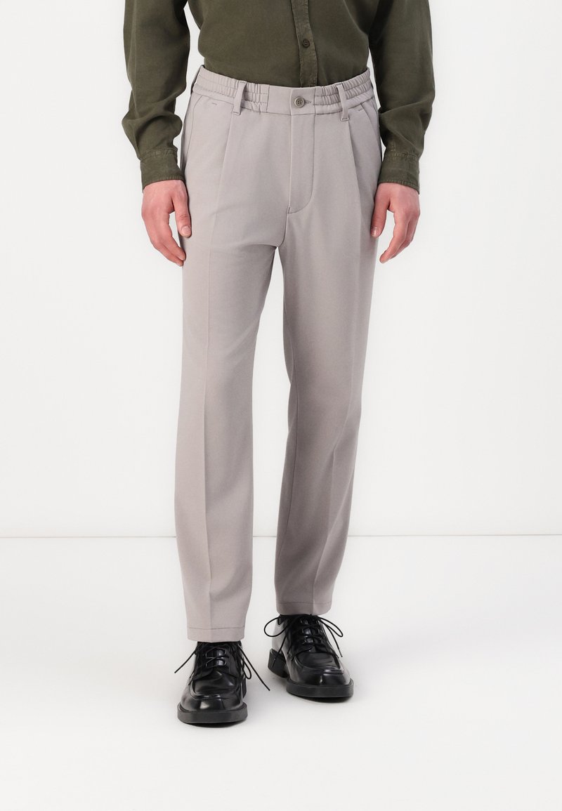 Pantalones ajustados de color gris claro con cintura elástica, pliegues frontales y bolsillos laterales; combinados con zapatos negros de cordones.