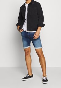 Man wearing rolled-up denim shorts, black jacket over white shirt, and black canvas sneakers standing against a plain background.