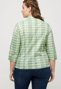 Camisa a cuadros verde y amarilla con mangas de tres cuartos, presenta un cuello alto y detalles en la costura trasera, combinada con pantalones de mezclilla oscuros.