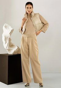 Vrouw in een beige gebreid sweater, puffer vest, wijde broek en laarzen met slangenprint poseert naast een abstract wit beeldhouwwerk op een zwart voetstuk.