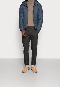 Giacca imbottita blu navy, dolcevita beige, pantaloni scuri e sneakers color cuoio. La giacca presenta un cappuccio, una texture liscia e una chiusura con zip.