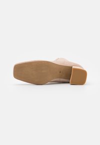Zapato de tacón bloque en ante beige con un diseño de punta redonda, que presenta una superficie lisa y ligeramente texturizada y una suela de caucho sólida.