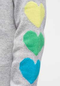 Μανίκι γκρι πλεκτού πουλόβερ με τρία χρωματιστά σχέδια καρδιάς σε κίτρινο, πράσινο και μπλε, τοποθετημένα κάθετα στον βραχίονα.