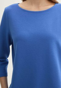 Blau strukturiertes Oberteil mit rundem Halsausschnitt und drei Viertel-Ärmeln, das ein geripptes Stoffmuster aufweist. Einfaches, elegantes Design.