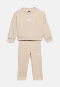 Bež dvoset trenirka z dolgimi rokavi in ujemajočimi hlačami, obe s belimi logotipi Adidas na prsih in stegnih.