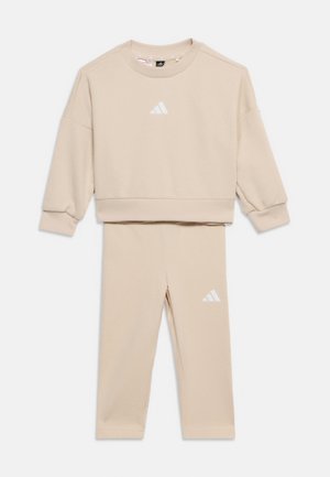 Beige trainingspak in tweeën, bestaande uit een sweatpant met lange mouwen en bijpassende broek, beide met witte Adidas-logo's op de borst en dij.