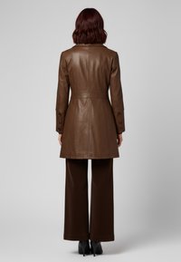 Bruine leren trenchcoat met een getailleerde taille, lange mouwen en met knopen gesloten manchetten. Gedragen over ruime bruine broeken. Gladde textuur.