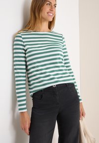 Camicia a maniche lunghe con righe orizzontali verdi e bianche, realizzata in materiale morbido. Abbinata a pantaloni neri e con una borsa di colore chiaro.