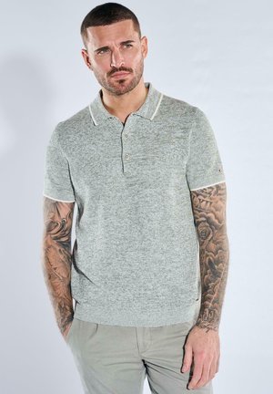 Mann mit kurzen dunklen Haaren und vollständig tätowierten Armen trägt ein hellgraues Poloshirt und helle Hosen, steht mit den Händen in den Taschen.