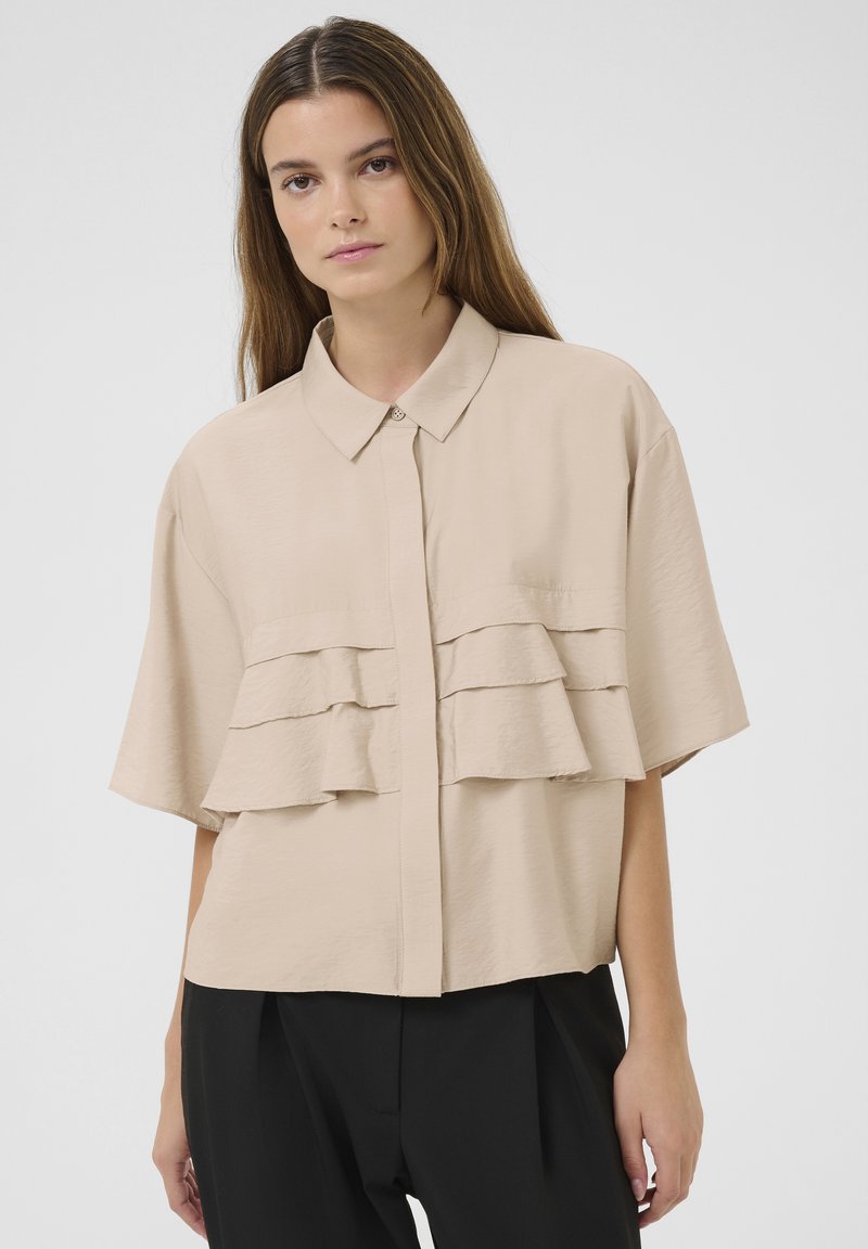 Short-sleeve beige shirt with layered ruffles, a collared neckline, and front button closure. Smooth fabric with a relaxed fit.