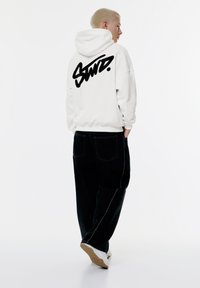 Vit huvtröja med svart grafisk text på baksidan, matchad med mörka jeans med kontrasterande sömmar, och ljusa sneakers.