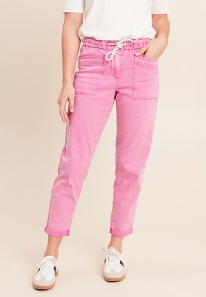 Person, die hochgekrempelte rosa Hose mit aufgesetzten Fronttaschen und weiße Turnschuhe mit schwarzen Streifen trägt, steht vor einem einfarbigen Hintergrund.