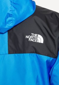 Blå och svart vattentät jacka med huva, med en texturerad yta och den vita logotypen för The North Face på det svarta området.