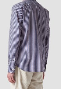 Hombre vistiendo una camisa de manga larga a rayas verticales en azul marino y blanco, con puños abotonados y pantalones de pana color crema, visto desde atrás.