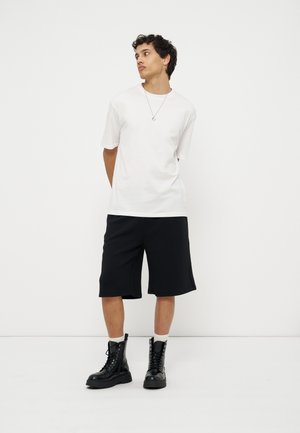 Mladý muž v bielom tričku, čiernych šortkách po kolená, čiernych šnurovacích topánkach a so náhrdelníkom, stojaci pred jednoduchou bielou stenou.