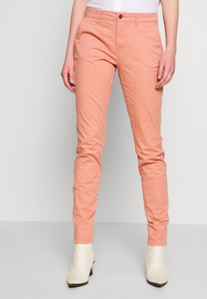 Person trägt eine eng anliegende, pfirsichfarbene Hose mit Knopf- und Reißverschluss, kombiniert mit weißen Stiefeletten.