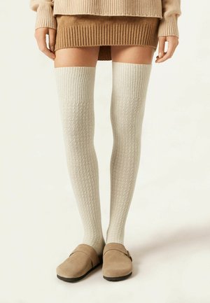 Chaussettes montantes couleur crème clair avec une texture côtelée et un motif géométrique, assorties à des sabots beiges à enfiler avec un détail de sangle.