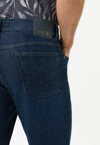 Mörkblå denimjeans med en bakficka med ett intrikat sömnadsmönster och en läderpatch med varumärke nära midjan.