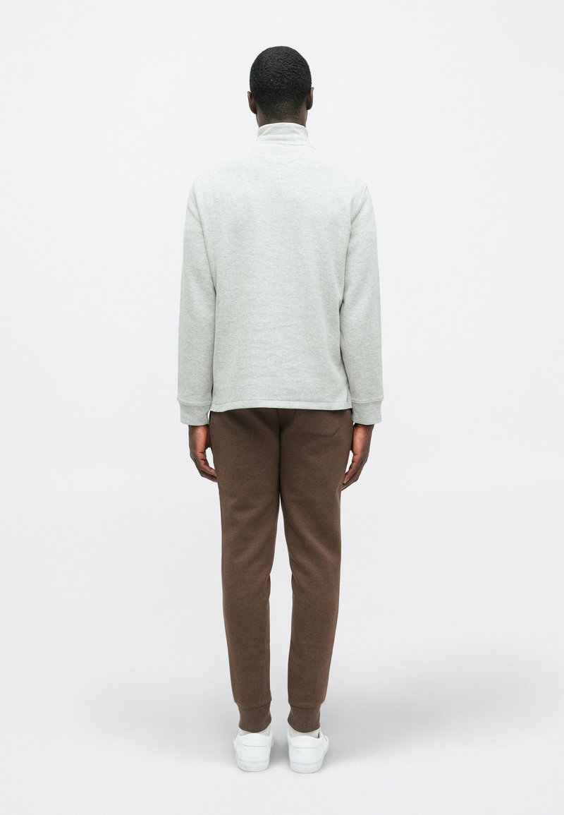 Grauer Pullover mit hohem Kragen, lockerer Passform und gerippten Bündchen, kombiniert mit braunen Jogginghosen und weißen Sneakern, von hinten betrachtet.