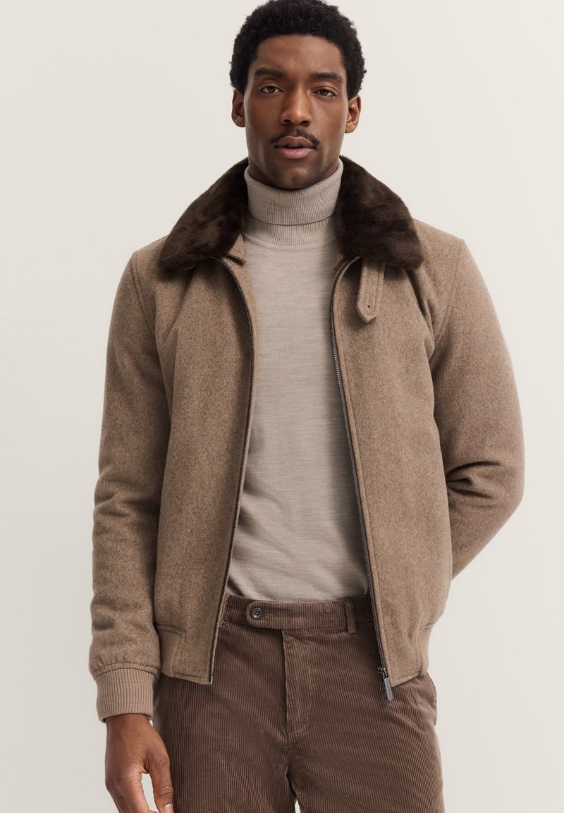 Brown wool blend jacket with a faux fur collar, front zipper, ribbed cuffs, worn over a light cream turtleneck and brown corduroy pants.