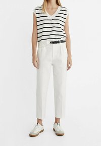 Gestreepte gebreide mouwloze top in crème en zwart, gecombineerd met hoge witte broeken en witte sneakers met beige zolen.