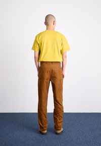 Tricou galben din bumbac purtat cu pantaloni din catifea fina maro. Croiala este relaxată, iar pantaloni au un eticheta mică cu logo pe buzunarul din spate.