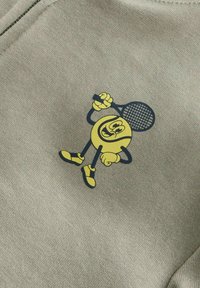 Lys grått stoff med en gul tegneseriefigur som holder en tennisracket. Designet inkluderer svarte konturer og lekne ansiktstrekk.