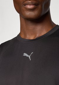 Svart atletisk T-skjorte med rund halslinning og en reflekterende grå Puma-logo på venstre brystområde. Glatt, tett tekstur.