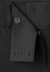 Prim-plan cu material negru care arată o clapetă cu nasturi parțial deschisă, având trei găuri verticale pentru nasturi și un singur nasture de plastic vizibil.