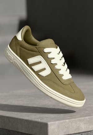 Zapatilla de gamuza verde con cordones y acentos blancos. Presenta una superficie texturizada y un logo. Diseño clásico de corte bajo, con suela de goma ligeramente elevada.