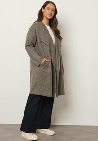 Cardigan lungo davanti aperto in morbido tessuto grigio, con due tasche laterali, vestibilità rilassata e maniche ampie, abbinato a jeans scuri e scarpe bianche.