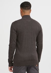 Dunkelbrauner gestrickter Pullover mit hohem Kragen, gerippten Bündchen und Saum, versehen mit Schulterpatches und einem strukturierten Muster auf dem Stoff.