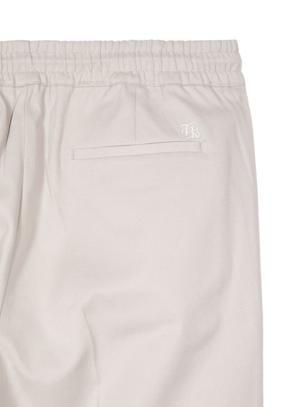Beige elastic waistband pants with a single back welt pocket and small embroidered initials near the pocket.