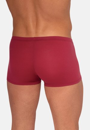 Rückenansicht einer Person, die eng anliegende, gerippte, dunkelrote Badehose vor einfachem Hintergrund trägt.