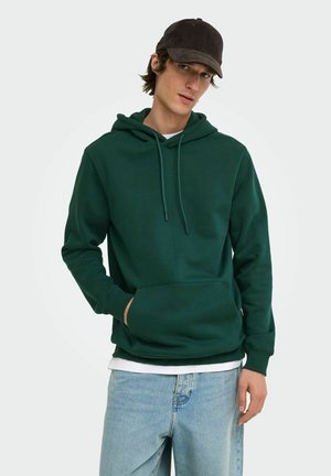 Junger Mann trägt einen dunkelgrünen Hoodie, hellblaue Jeans und eine schwarze Baseballkappe, steht mit einer Hand in der Tasche.