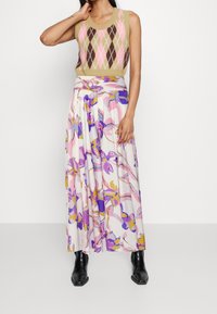 Kobieta wearing a bezrękawnik w wielokolorowy wzór argyle w odcieniach beżu, różu i brązu, zestawiony z kwiecistą spódnicą maxi w kolorach purpurowym, różowym i żółtym oraz czarnymi butami.