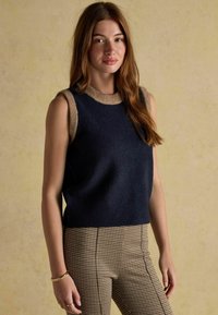 Navy sleeveless sweater with a beige round neck and armhole borders, featuring a soft texture. Paired with houndstooth patterned pants.