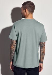 Světle zelené tričko s krátkým rukávem, kulatým výstřihem, měkkou texturou a uvolněným střihem. Viditelné vzory tetování na ruce.