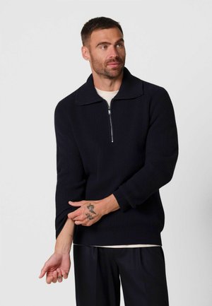 Man past mouw van een zwarte geribbelde trui met kwart-ritssluiting aan, draagt zwarte broek en licht T-shirt, met zichtbare handtattoos.